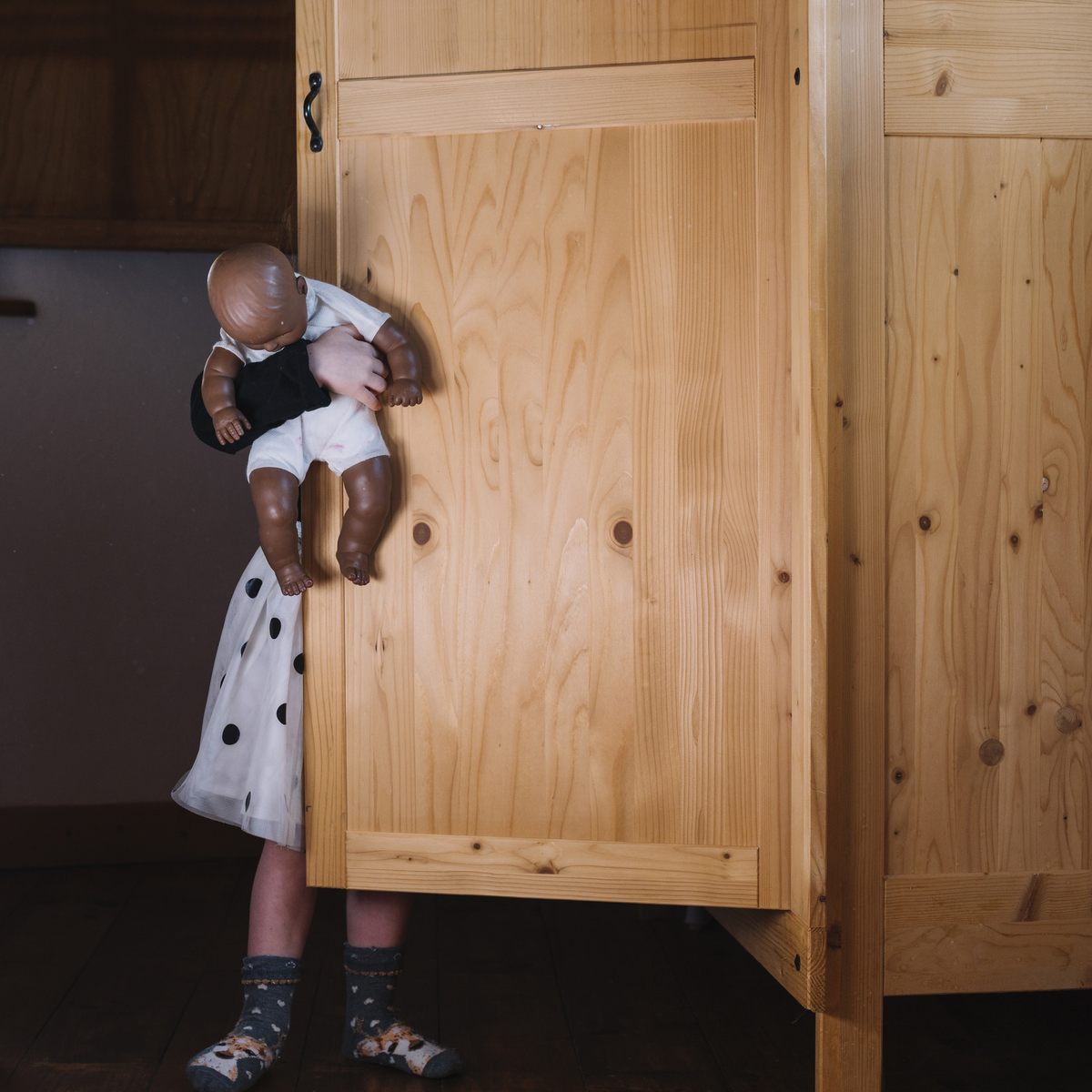 Ein Kind versteckt sich mit Puppe hinter einem Holzschrank.