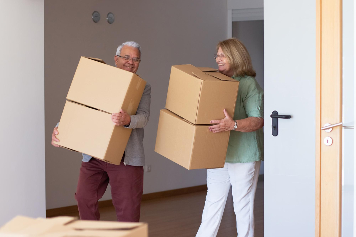 Älteres Paar trägt lächelnd Umzugskartons in neue Wohnung.