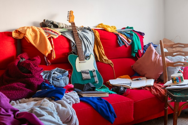 Unordentliches rotes Sofa mit Kleidung und E-Gitarre.